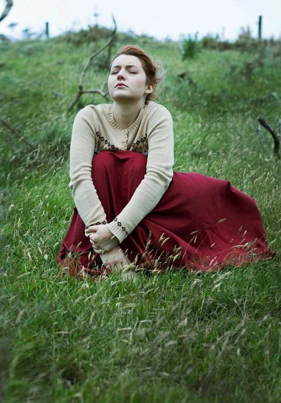 A woman with closed eyes sitting on grass in a field, wearing a beige sweater and a long red skirt.
