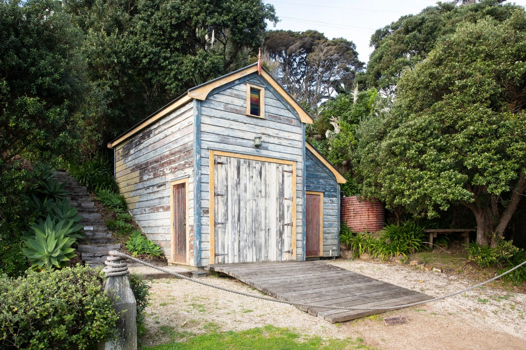 A weathered wooden shed at Little Oneroa Beach on Waiheke Island surrounded by lush green trees and plants, with a small wooden pathway leading up to it and a rope barrier in front.