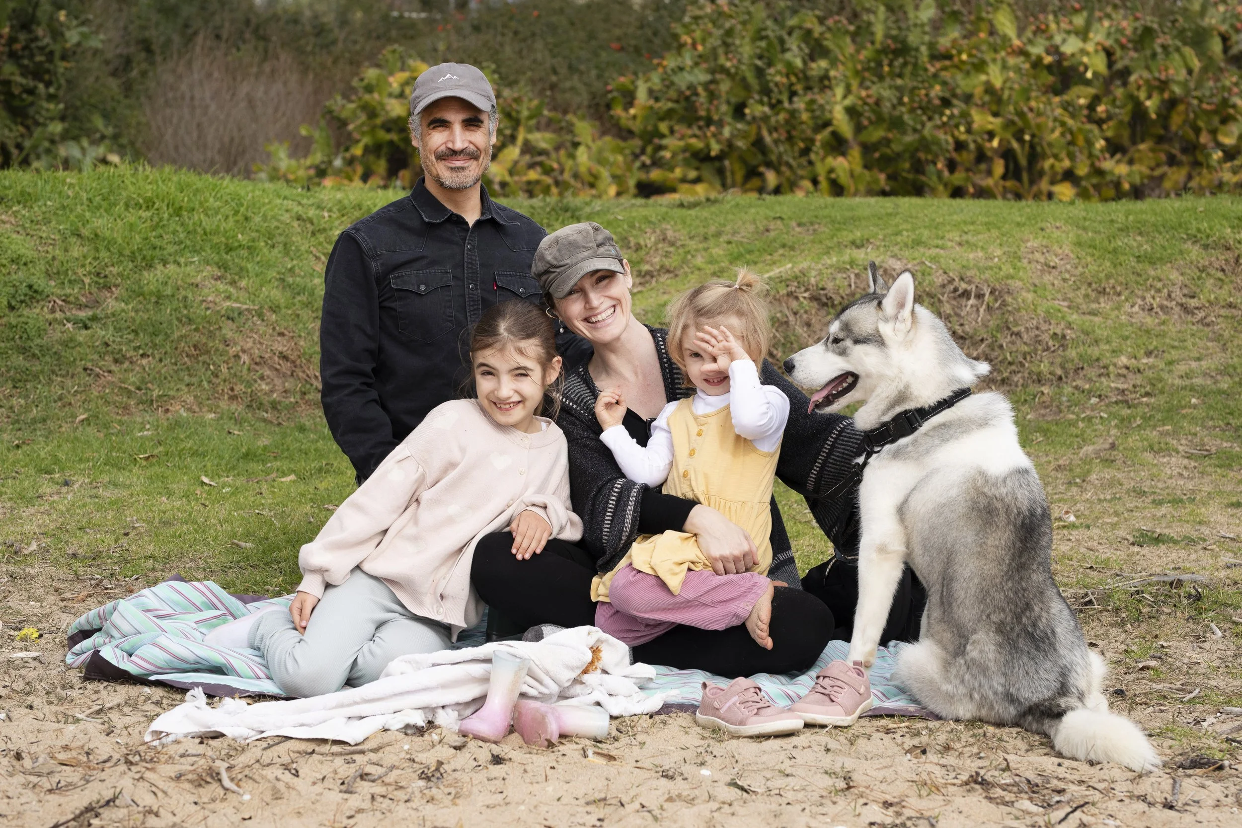 A group of five people, including two adults and three children, sitting on a blanket outdoors with a Siberian Husky dog, in front of a grassy area and bushes.