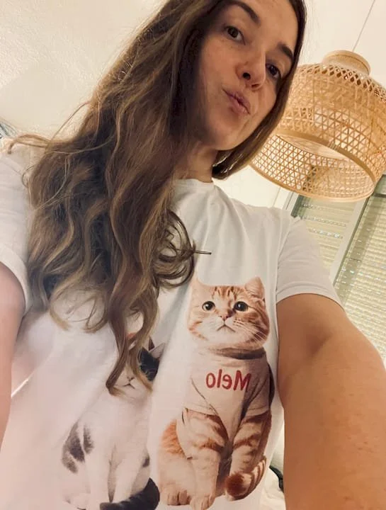 A woman taking a selfie, wearing a white T-shirt with a picture of a cat on it. The cat on the shirt has a serious expression and is wearing a sweatshirt with the word 'Melo' on it. There is a woven lamp hanging from the ceiling behind her.