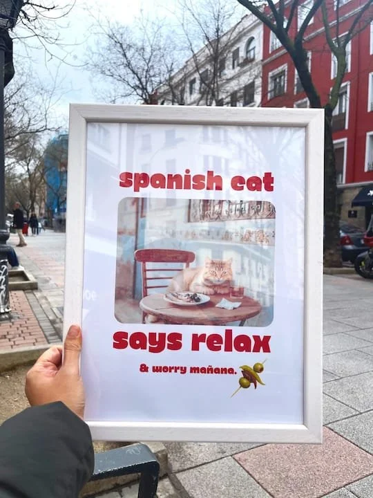Sign advertising a Spanish cat; the sign shows a photo of a cat sitting at a table with a plate, and text that reads 'Spanish cat says relax & worry mañaña' with a small illustration of a pintxo gilda.