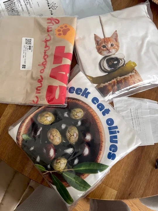 Photo of a t-shirt samples one with black and green olive with cat faces, a T-shirt with a cat face on top of a spanish tapa, and a tote bag with the words ask me about my cat on a wooden table.
