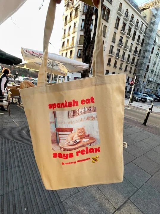 A beige tote bag hanging outside on a city street, with a picture of a cat sitting at a table eating, and red text that reads "spanish cat says relax" and smaller text "& worry mañana."