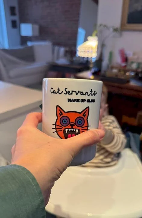 A person holding a ceramic mug with the text 'Cat Servants Wake Up Club' and an illustration of an orange cat with purple eyes and sharp teeth.