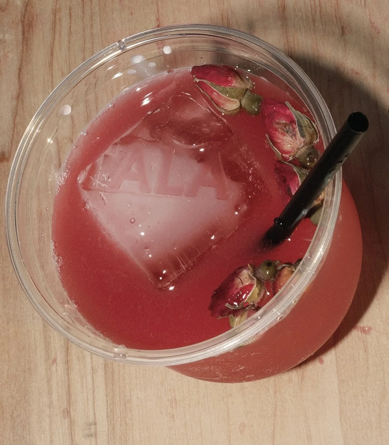 A clear plastic cup filled with pink-colored drink, ice cube, rosebuds, and a black straw, placed on a wooden surface.