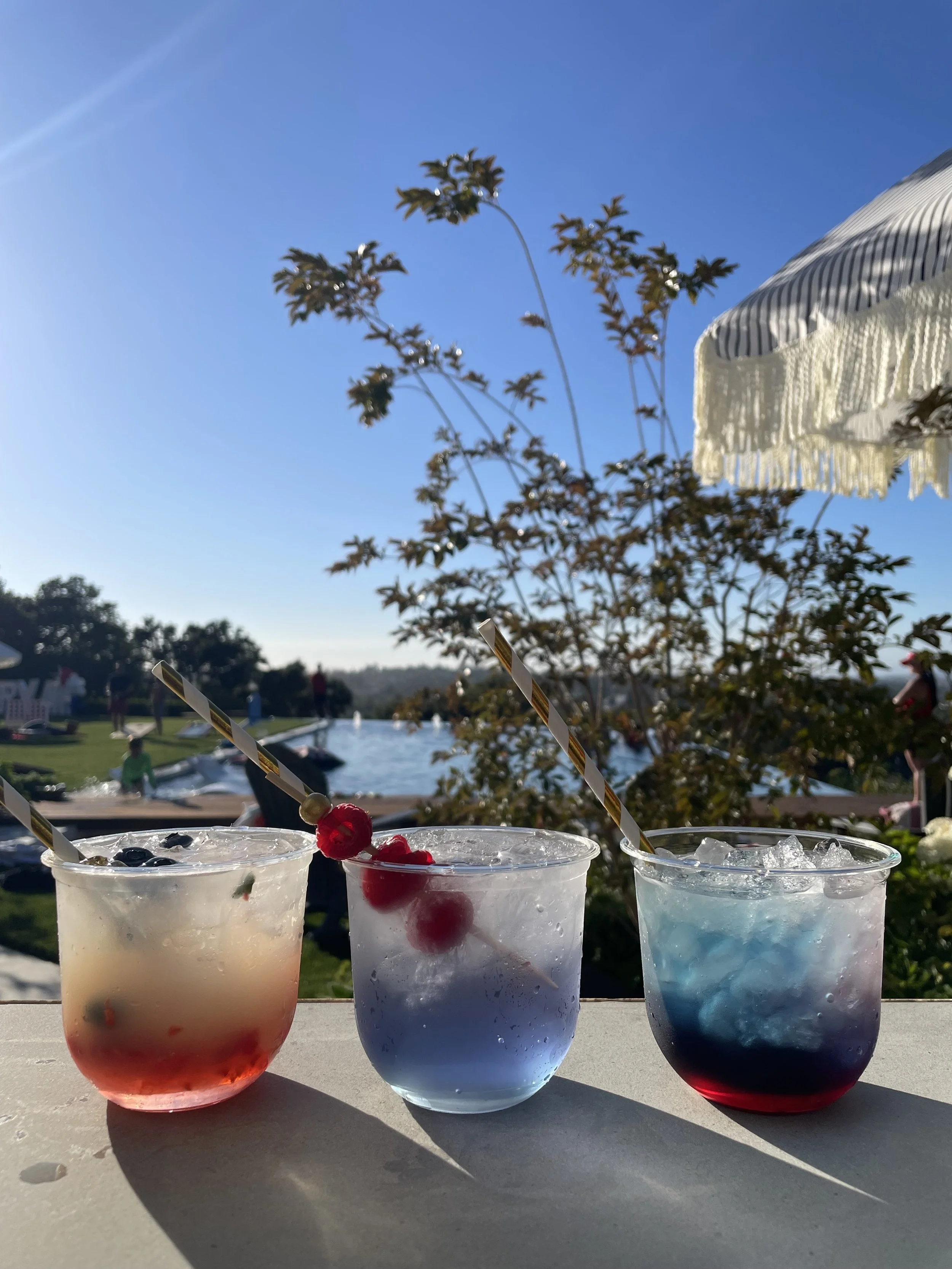 Three colorful cocktails with ice on a table outdoors, with a pool, trees, and a clear blue sky in the background, under a striped umbrella.