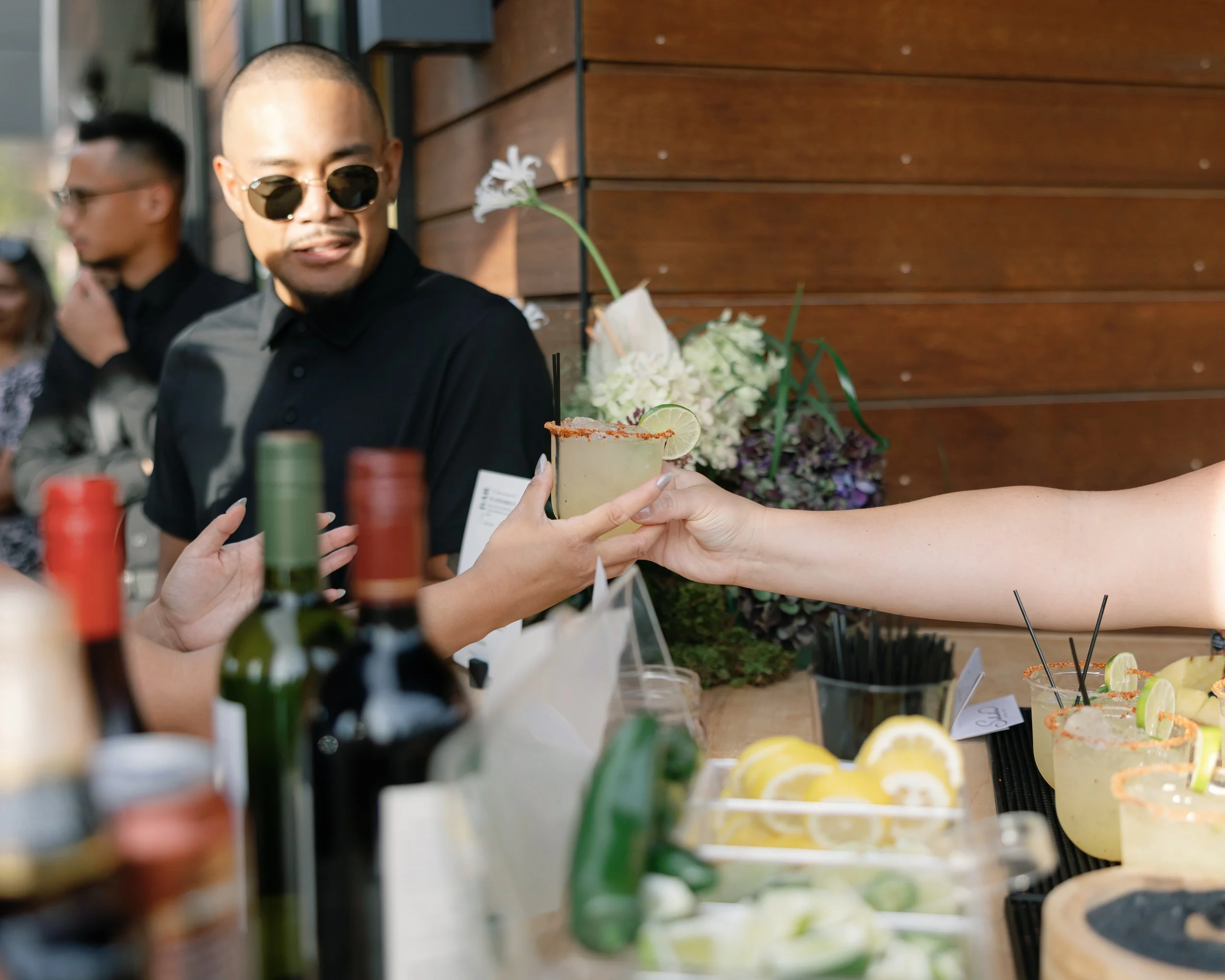 Person in black shirt and sunglasses reaching for a margarita with a lime wedge at a social gathering, with others and bottles visible on the table.