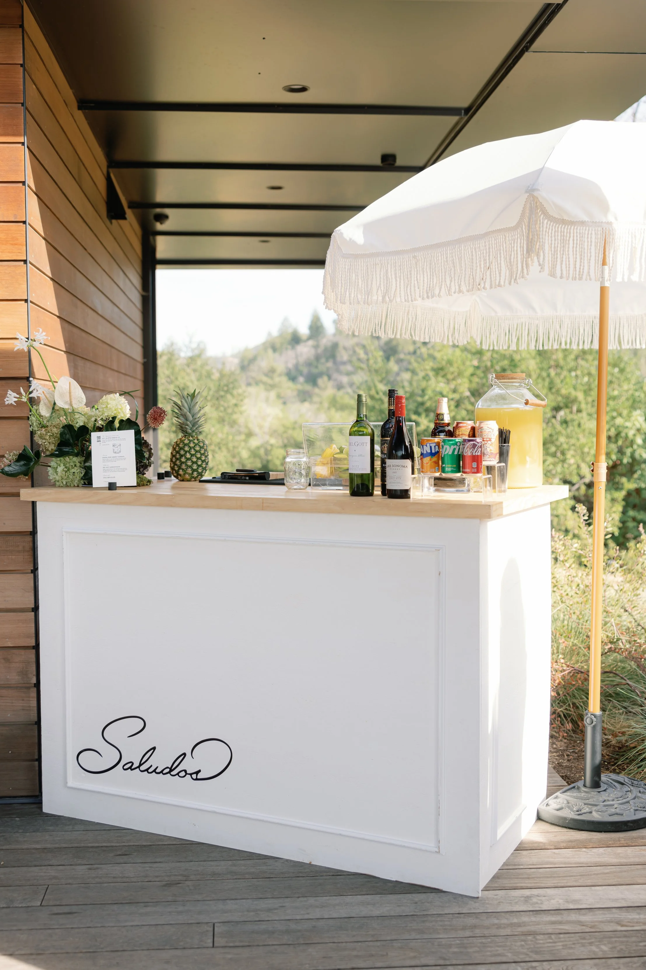 Outdoor bar cart with drinks, glassware, pineapple, flowers, and a white umbrella on a wooden deck with a scenic mountain and trees background.
