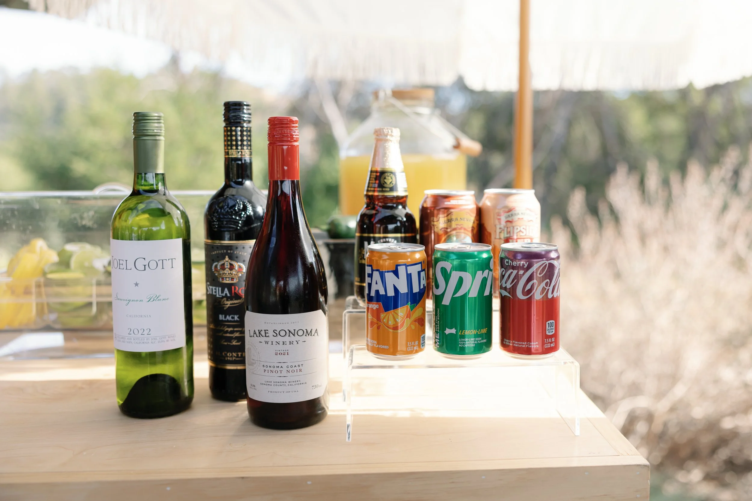 Assorted drinks including a bottle of white wine, a bottle of red wine, two cans of soda (Fanta, Sprite, Cherry Cola), a bottle of beer, and a can of beer, set on a wooden table with a blurred outdoor background.
