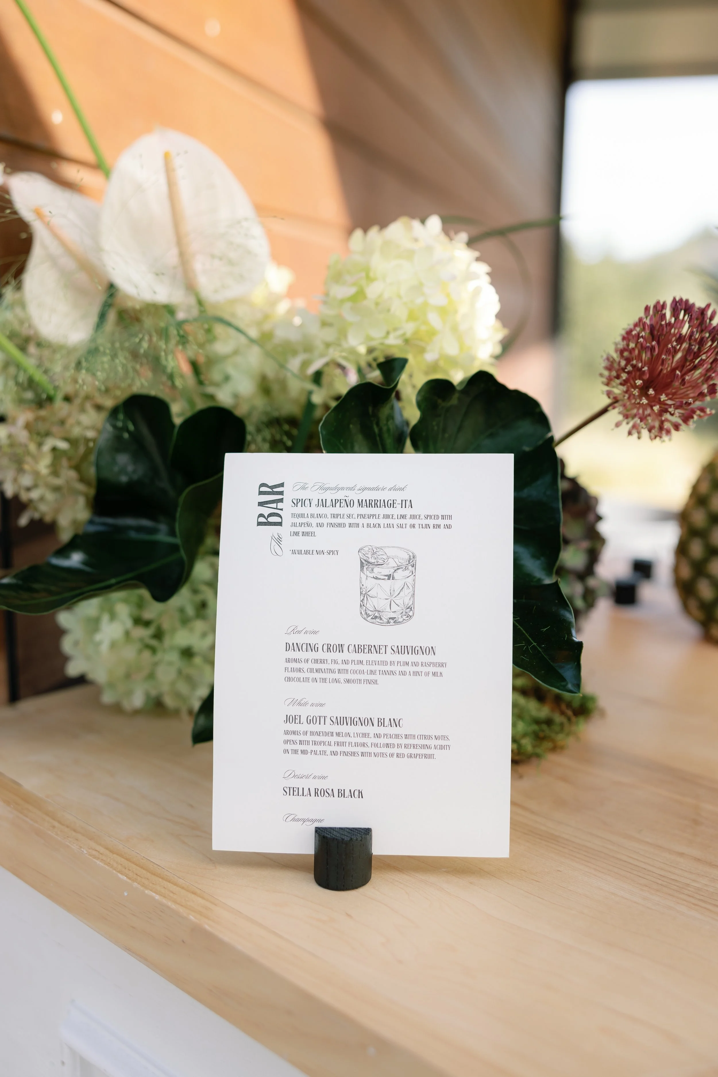 Event menu card featuring drinks and food options, displayed on a table with floral arrangements in the background.