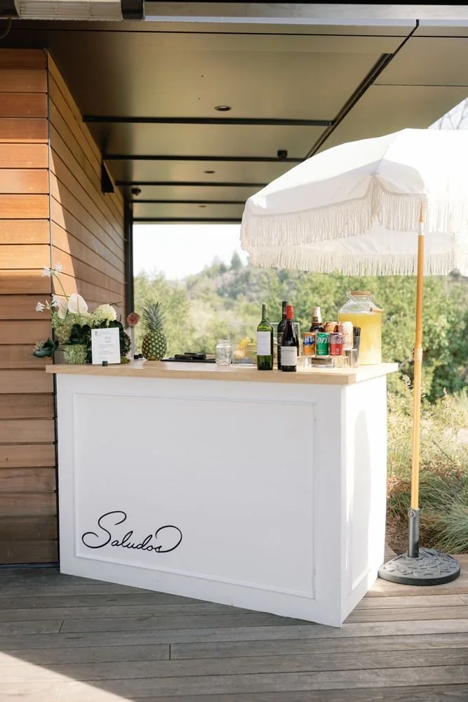 A small outdoor bar with a white counter with the word 'Saludos' written on it, topped with bottles of wine and soda, a pineapple, flowers, and a large white umbrella providing shade, set against a natural landscape background.