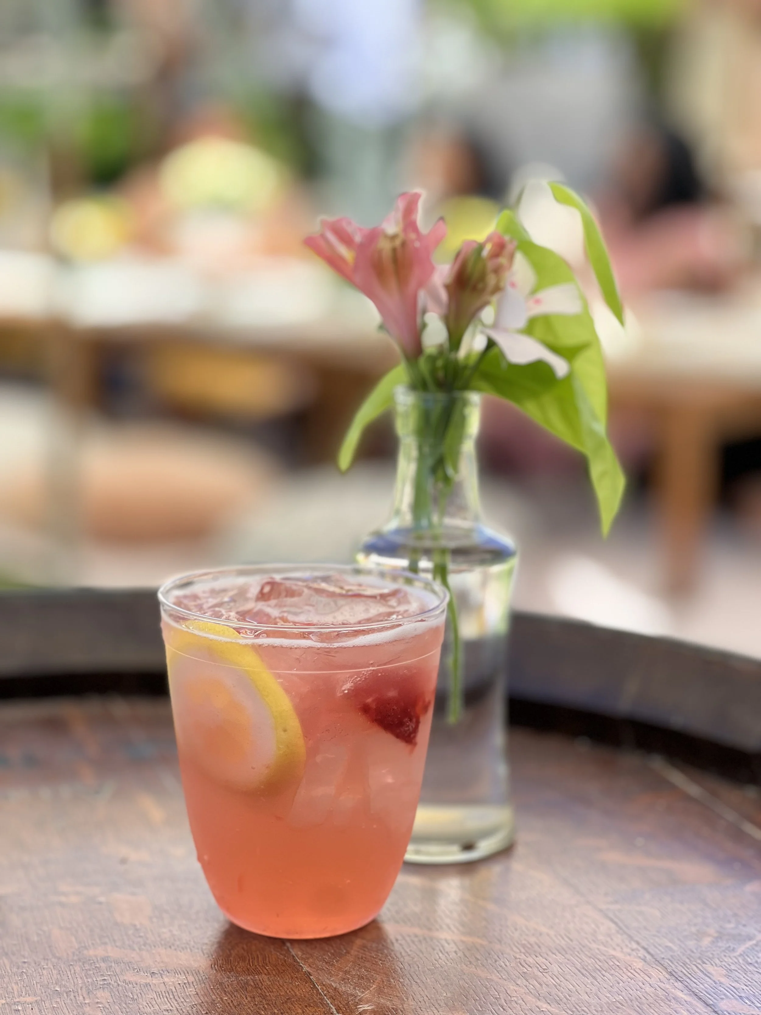 A pink-colored drink with lemon slices and berries on a wooden table, with a vase of pink flowers in the background.