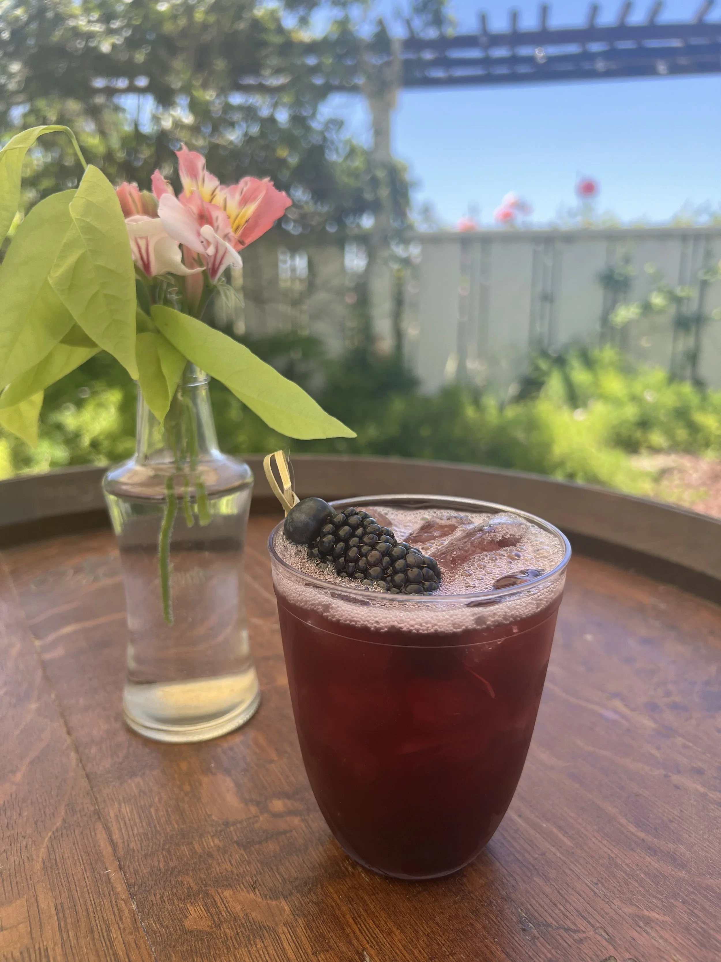 A glass of dark purple berry beverage with a blackberry garnish on ice, placed on a wooden table outdoors with a blooming flower arrangement in a small glass vase in the background and a fence with trees and sky beyond.