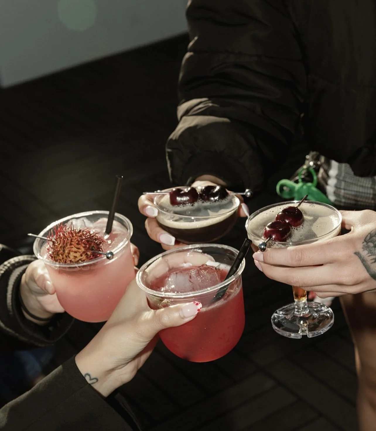 Four people holding cocktails, three with pink and red drinks with cherries or fruit garnishes, and one with a dark drink garnished with cherries.