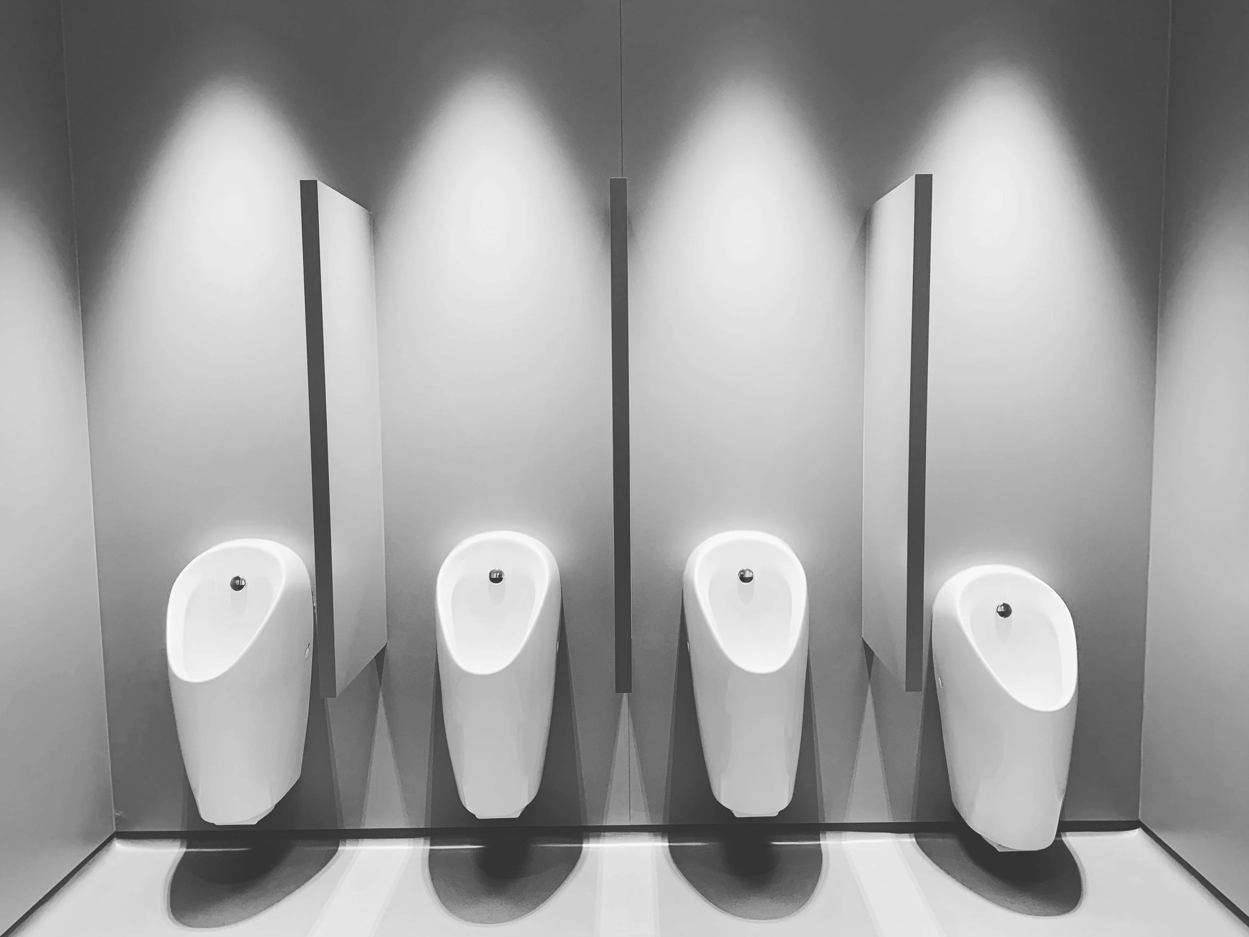 Six white urinals mounted on a gray wall with bright overhead lights in a restroom.