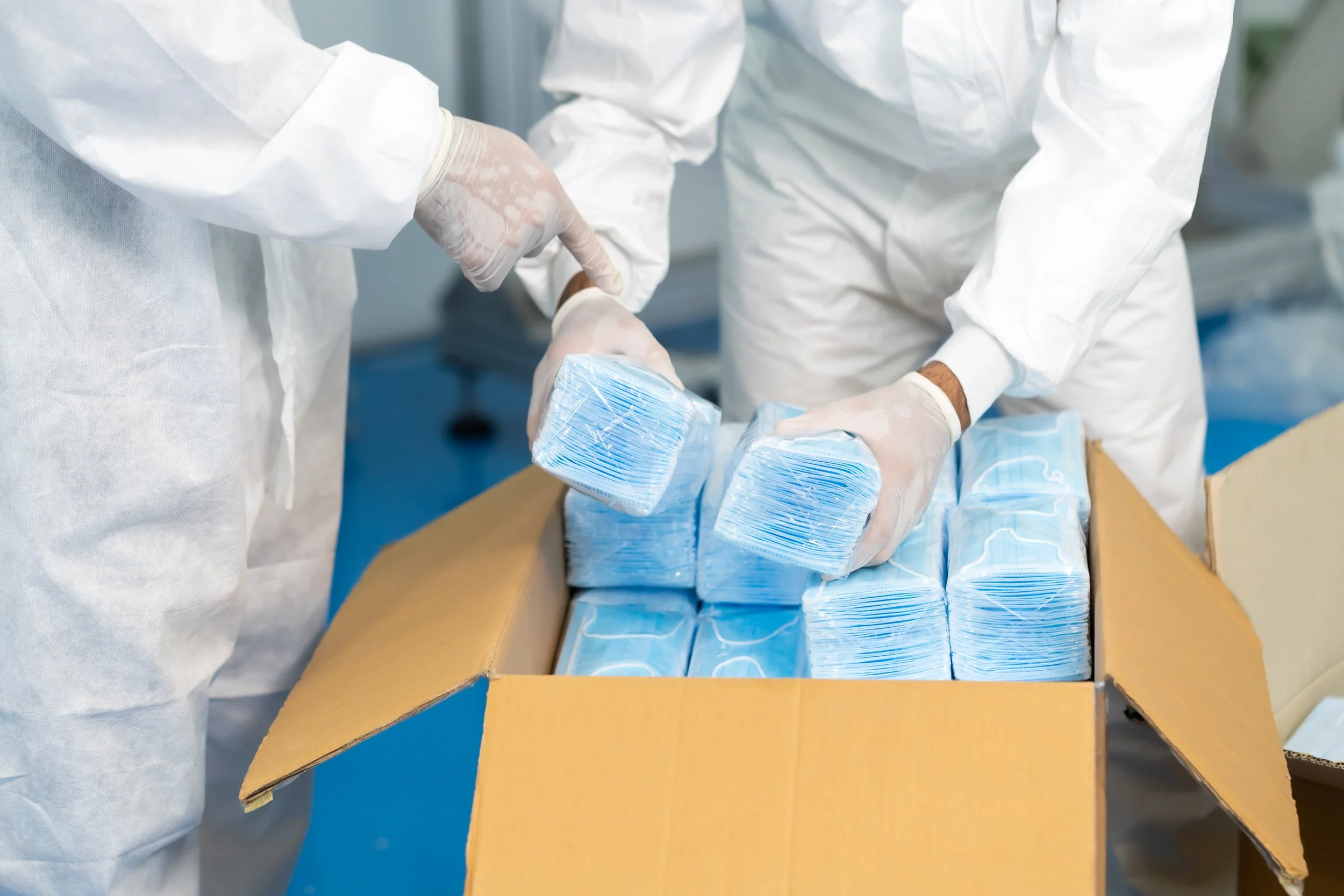 Two individuals in white protective suits and gloves pack blue and white face masks into a cardboard box on a table.