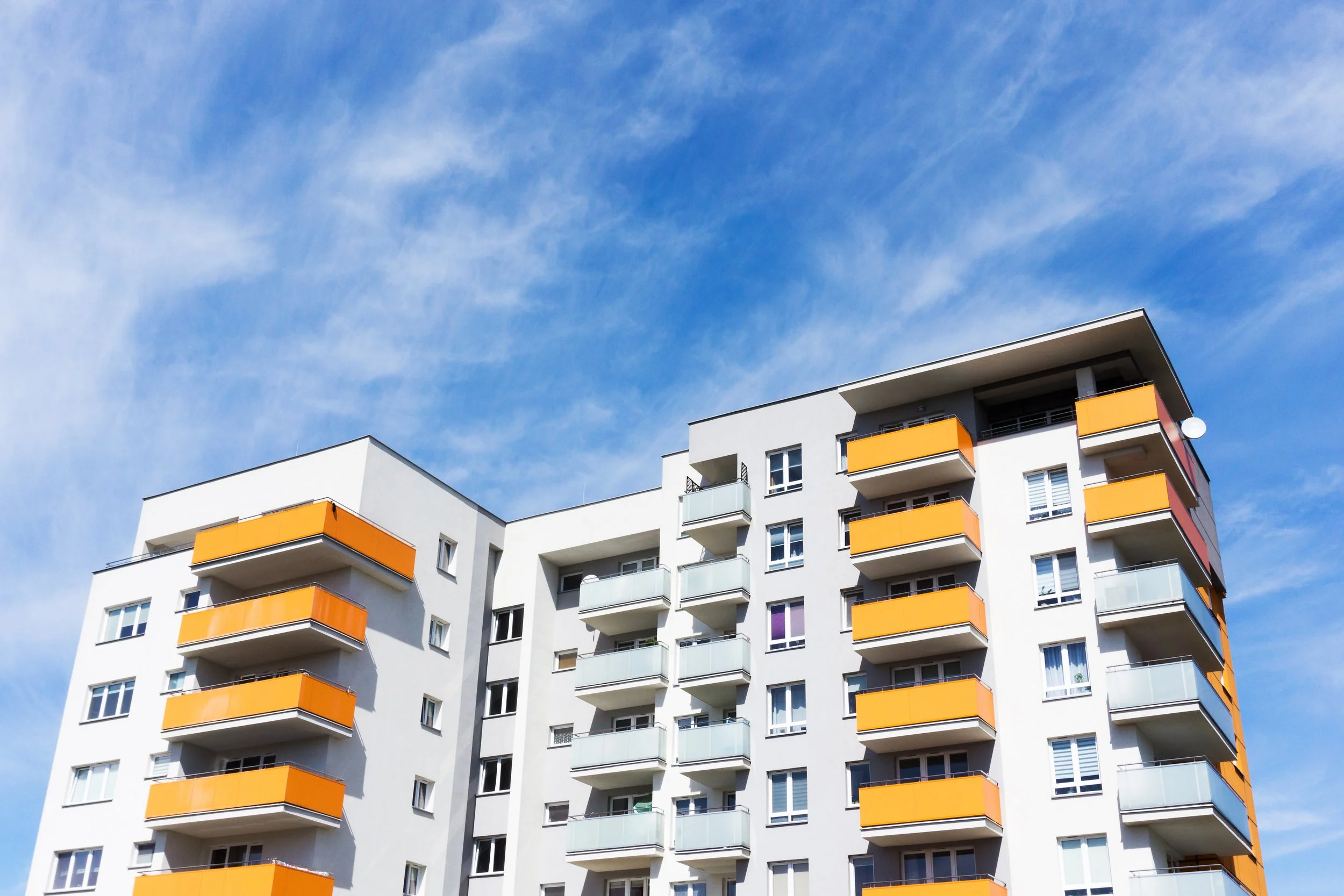 Modern white apartment building with orange balconies against a bright blue sky.