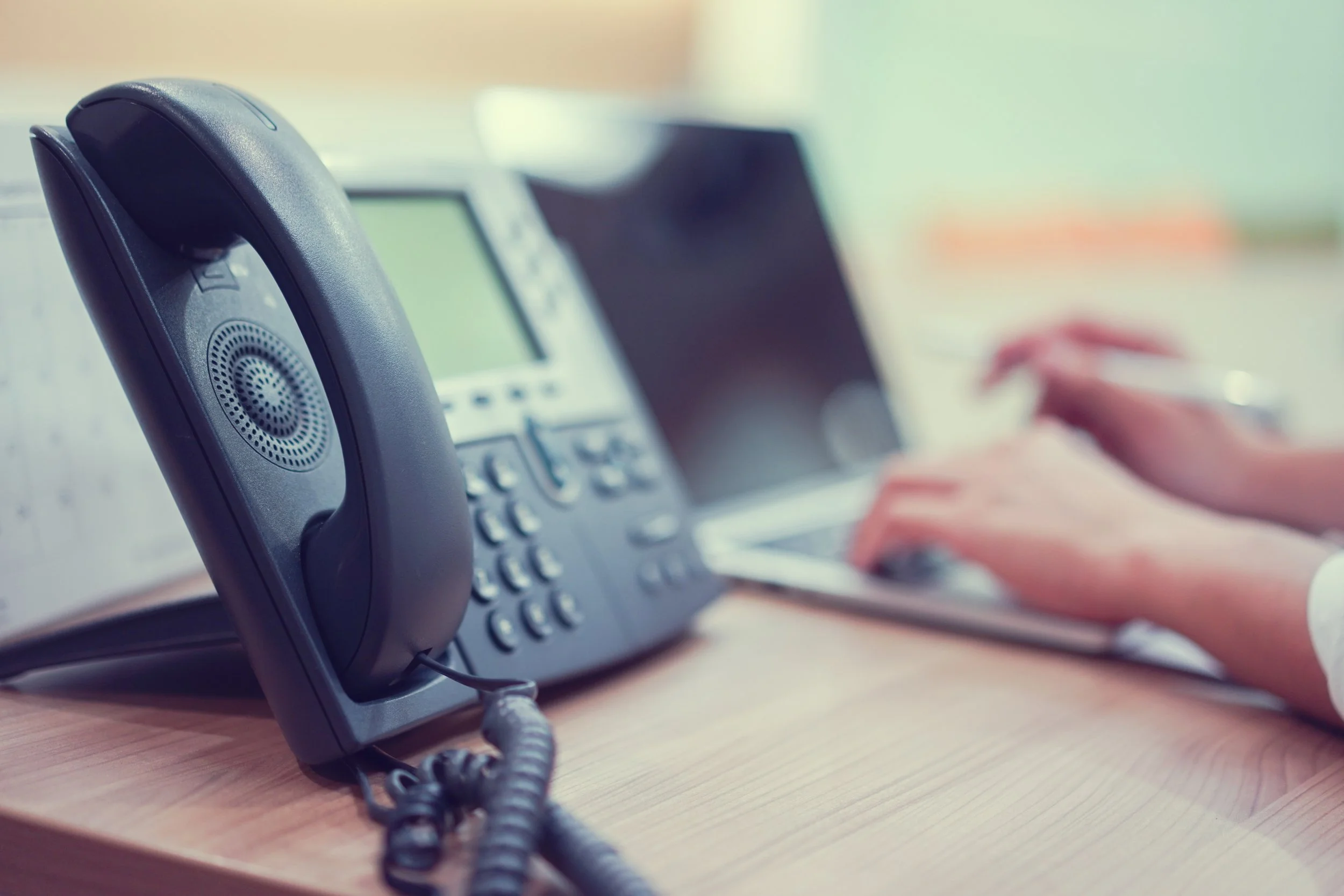 Close-up of a black office desk phone with a person's hands typing on a keyboard in the background at a desk.
