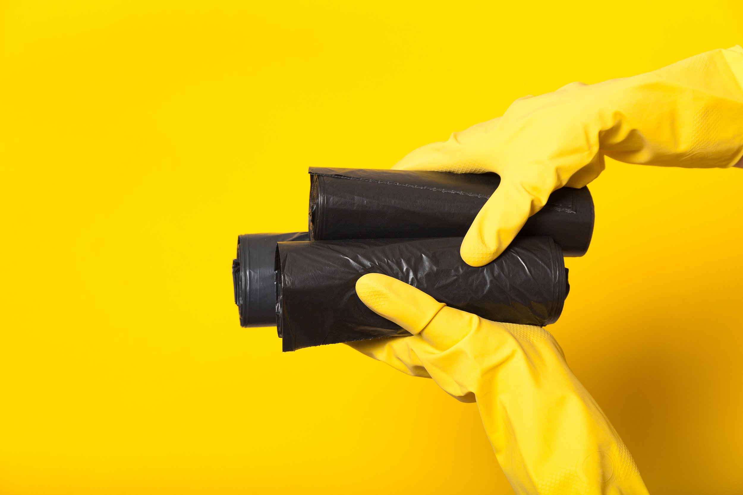 A hand wearing a yellow rubber glove holding three black garbage bags against a yellow background.