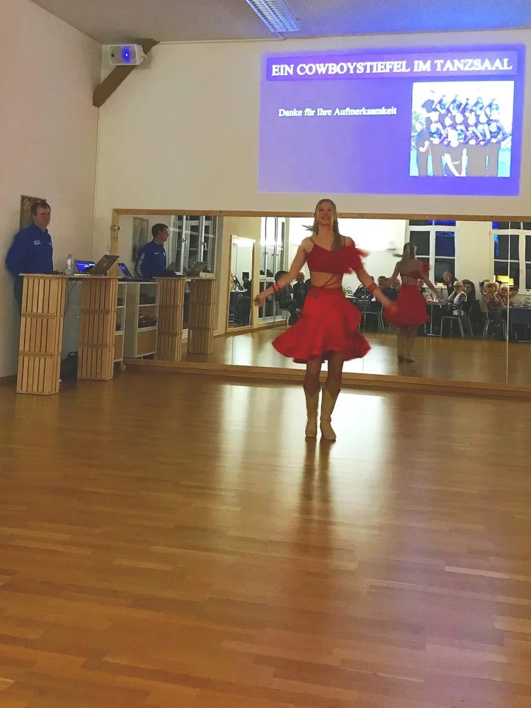 Tänzerin in rotem Kleid tanzt im Tanzsaal, im Hintergrund eine Projektionsleinwand mit der Aufschrift 'Ein Cowboy-Stiefel im Tanzsaal' und einer Dankesnachricht, links stehen drei DJs.