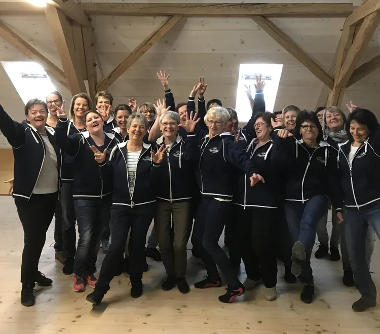 Gruppe älterer Frauen in dunkelblauen Jacken in einem Holzraum, die fröhlich lachen und feiern.