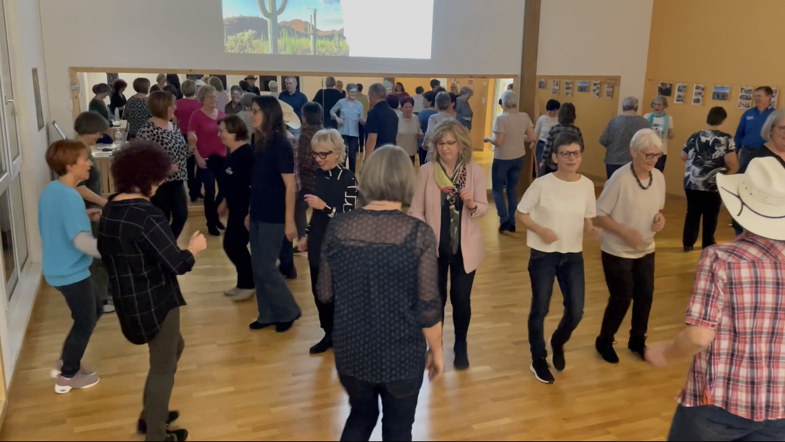 Menschen, die in einem Raum tanzen und sich unterhalten, mit einer Wand im Hintergrund, auf der Fotos und ein Bild mit Kakteen an der Wand hängen.