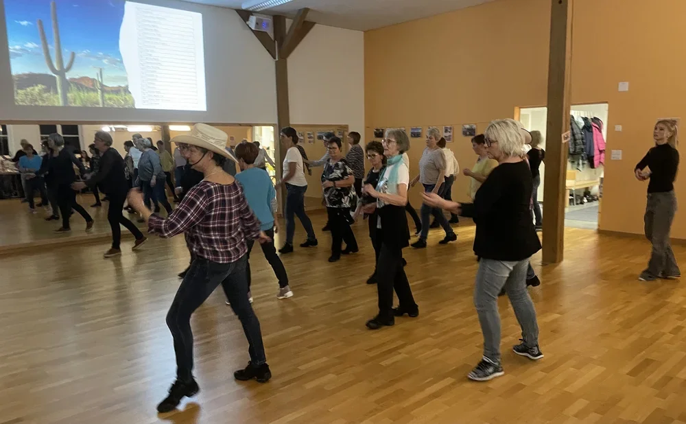 Menschen tanzen in einem Saal, einige im Western- oder Country-Stil, mit Holzfußboden, eine Frau trägt einen großen Sonnenhut, im Hintergrund eine Projektionsfläche mit einem desertähnlichen Bild.
