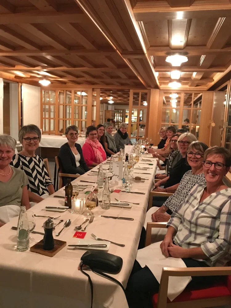 Eine Gruppe Frauen sitzt an einem langen gedeckten Esstisch in einem Holzraum und genießt ein gemeinsames Abendessen.