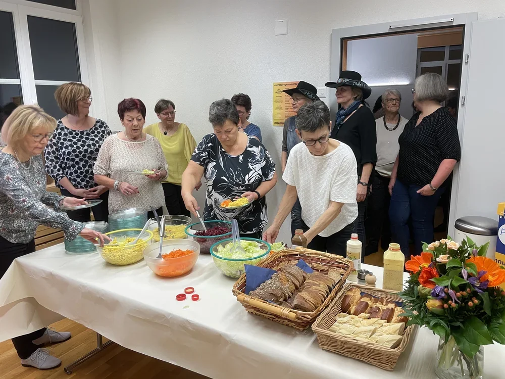 Menschen beim Buffet mit Salaten und belegten Broten in einem Gemeinschaftsraum.