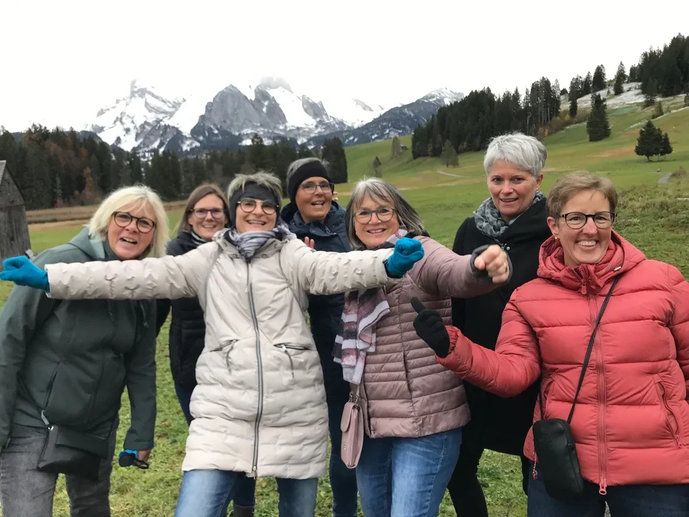 Eine Gruppe von sieben Frauen, die in einer grünen, bergigen Landschaft mit schneebedeckten Bergen im Hintergrund Spaß haben.