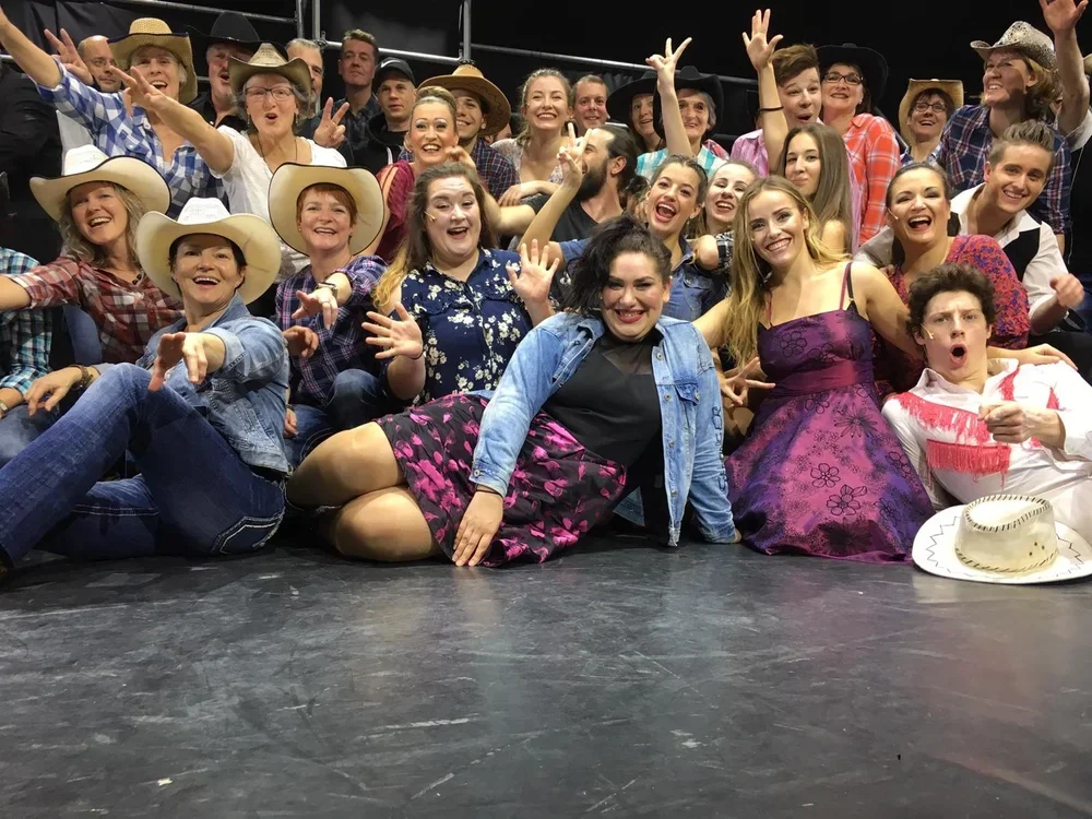 Gruppe von Menschen, die auf einer Bühne lachen, winken und feiern, viele tragen Cowboyhüte und Westernkleidung, einige sitzen auf dem Boden, bei einem freundlichen, lebhaften Gruppenfoto.