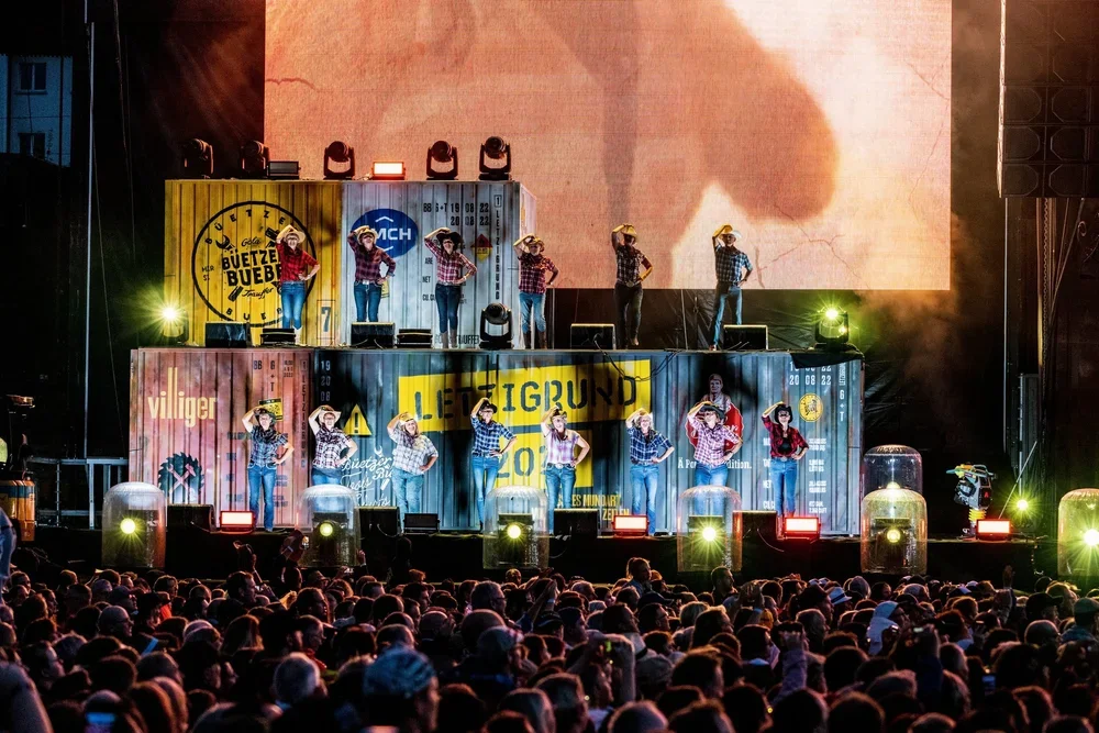 Eine Bühne mit mehreren Tänzern in Cowboystiefeln und Hüten, vor einer großen Zuschauermenge. Es gibt bunte Container-Wände im Hintergrund und Licht- und Bühneneffekte.
