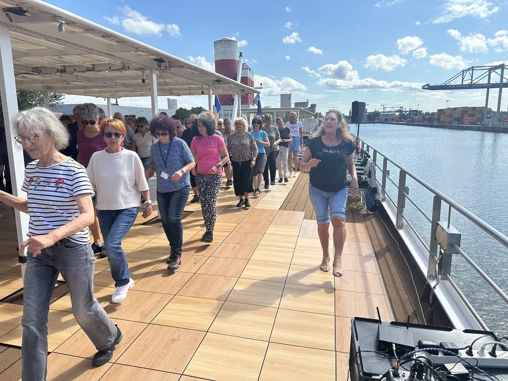 Menschen tanzen auf einem Deck neben einem Wasserweg an einem sonnigen Tag.
