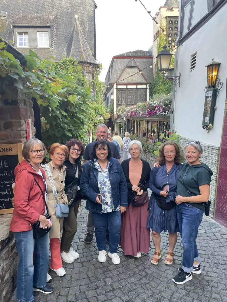 Eine Gruppe von acht Menschen steht in einer engen Gasse mit Kopfsteinpflaster, umgeben von bunten Gebäuden und blühenden Pflanzen. Es ist wahrscheinlich eine touristische oder Freizeit-aktivität in einer europäischen Stadt.