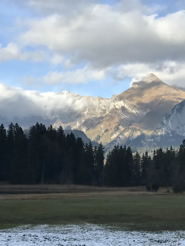 Berglandschaft mit schneebedeckten Gipfeln, Wolken am Himmel, eine Baumgruppe im Vordergrund und ein Gebiet mit Wasser oder Schnee am Boden.