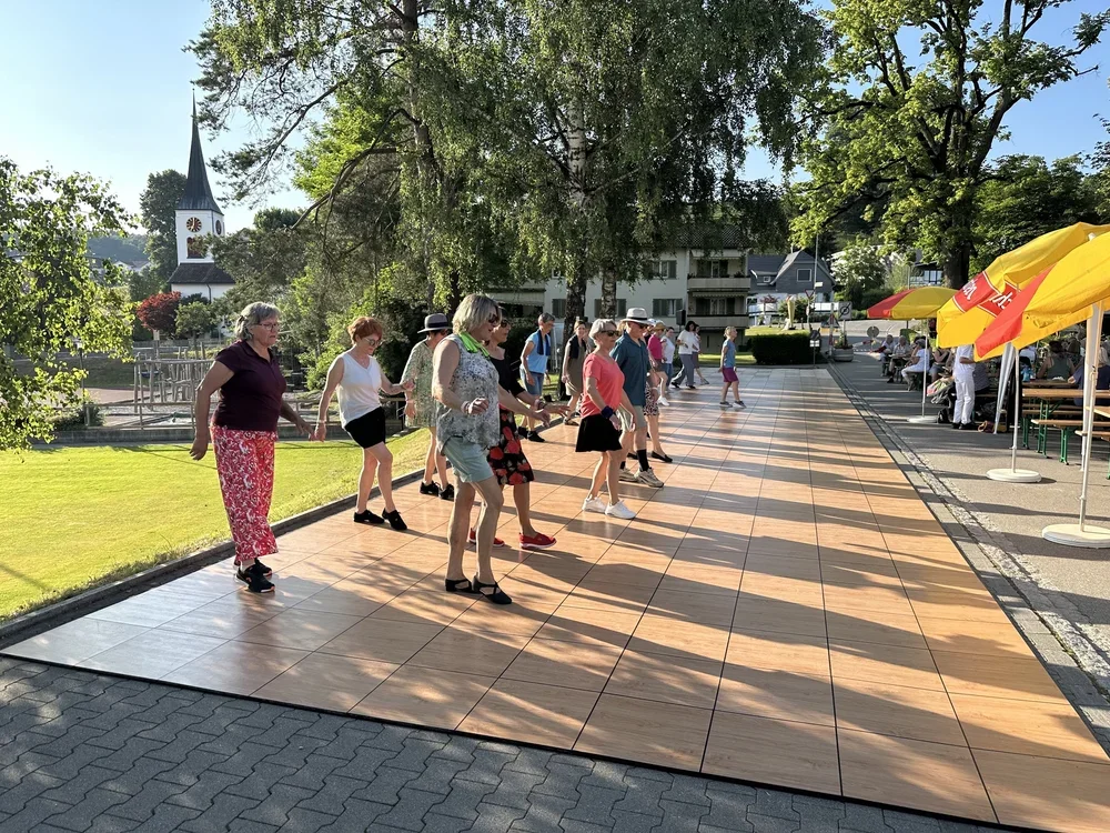 Menschen beim Tanzen auf einer Tanzfläche im Freien an einem sonnigen Tag, mit Bäumen im Hintergrund und einem Kirchturm sichtbar.