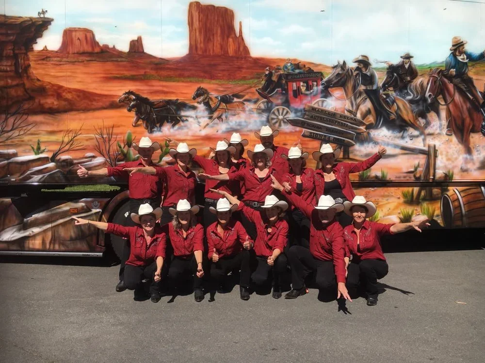 Gruppen von Menschen in Cowboy-Kostümen stehen vor einem Wandbild, das eine Western-Szene mit Pferden in der Wüste zeigt.