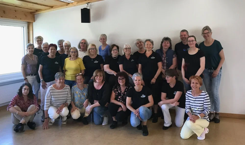 Gruppe von ca. 20 Menschen, hauptsächlich Frauen, in einem hellen Raum, einige tragen schwarze T-Shirts mit einem Logo, alle posieren für ein Gruppenfoto.