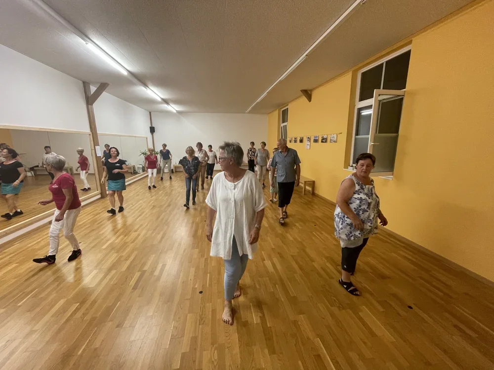 Mehrere Frauen tanzen in einem Raum mit Holzfußboden, weiße und gelbe Wände, einige haben kurze Haare, tragen legere Kleidung und tanzen gemeinsam.