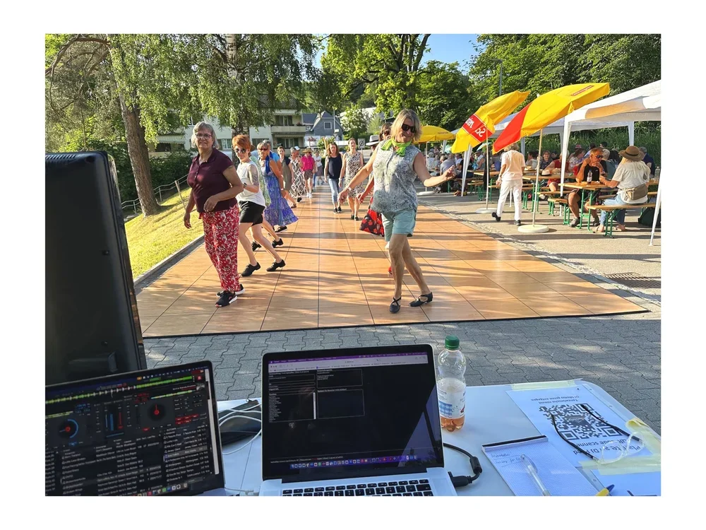Menschen tanzen im Freien auf einer Tanzfläche, im Hintergrund sitzen Leute an Tischen unter gelben Sonnenschirmen, es ist sonnig und es gibt viele Bäume.