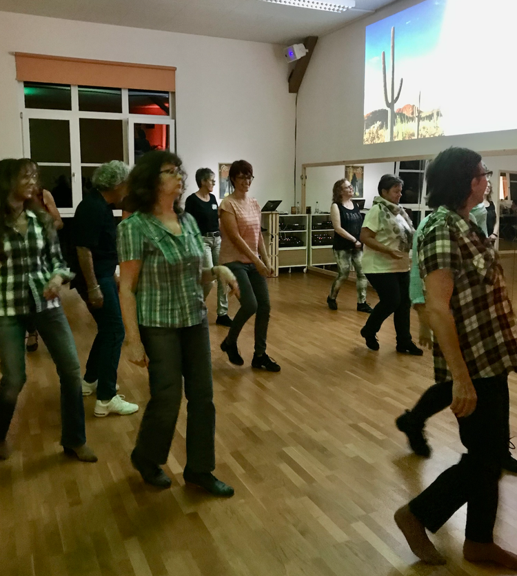 Mehrere ältere Frauen tanzen in einem Raum mit Holzboden, während im Hintergrund ein Bildschirm eine Wüstenlandschaft mit Kakteen zeigt.