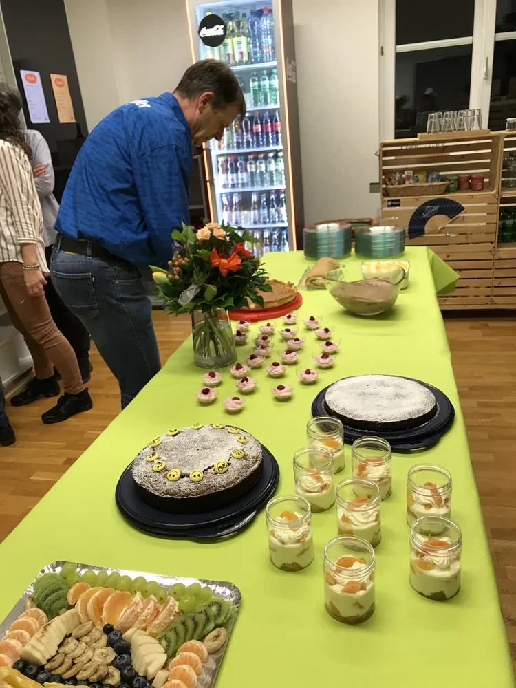 Tisch mit verschiedenen Desserts und Früchten, Blumenstrauß in der Mitte, Getränke im Kühlschrank im Hintergrund.