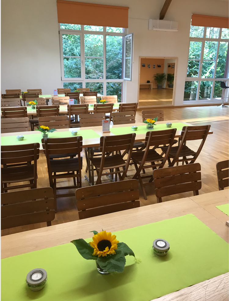 Ein heller Gartenraum mit großen Fenstern, mehreren Holzstühlen und Tischen, die mit hellgrünen Tischläufern, Sonnenblumen in Vasen und kleinen Kerzen dekoriert sind.