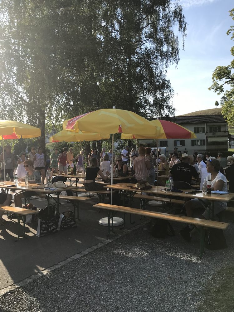 Menschen sitzen und stehen bei einem Treffen im Freien, mit bunten Sonnenschirmen, Bäumen und Gebäuden im Hintergrund.