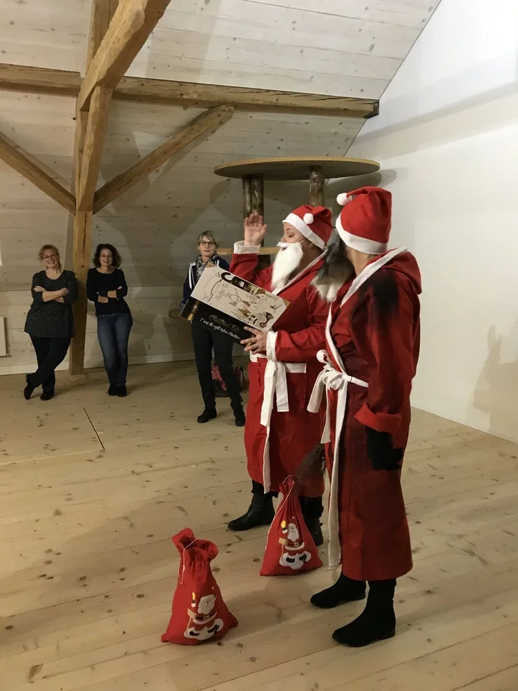 Zwei Personen in Weihnachtskostümen als Weihnachtsmann, die einen Geschenksack halten, während sie von mehreren Leuten amüsiert beobachtet werden.