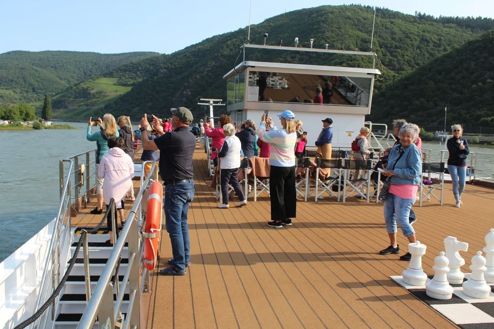 Menschen auf einem Schiff beim Fotografieren, mit Blick auf einen Fluss und grüne Hügel im Hintergrund.