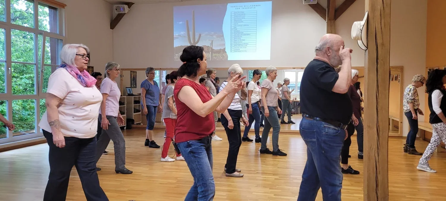 Ältere Menschen, die an einem Tanzkurs in einem hellen Raum mit großen Fenstern teilnehmen. Sie bewegen sich im Takt und haben Spaß. Auf der Wand ist ein Bildschirm mit einer Willkommensnachricht und einer Liste. Es herrscht eine freundliche Atmosphä