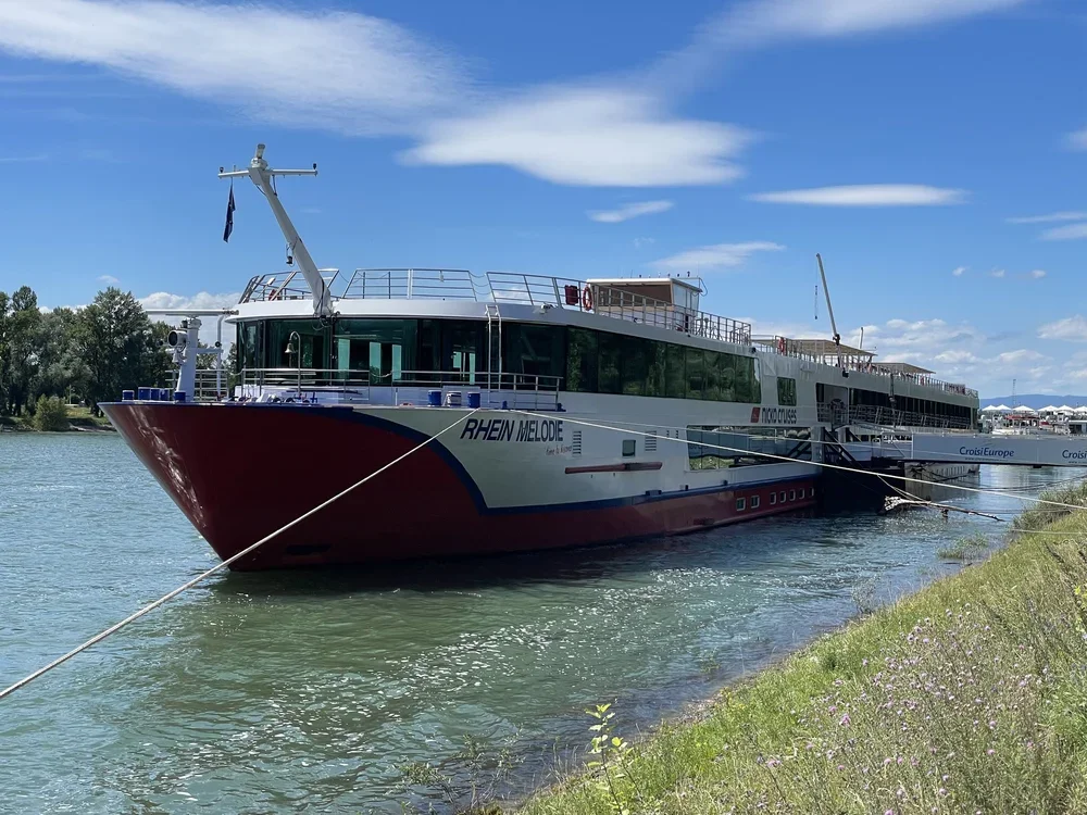 Großer Flusskreuzfahrtschiff mit dem Namen 'RHEIN MELODE', das am Ufer liegt, bei schönem Wetter mit blauer Himmel und einigen Wolken.