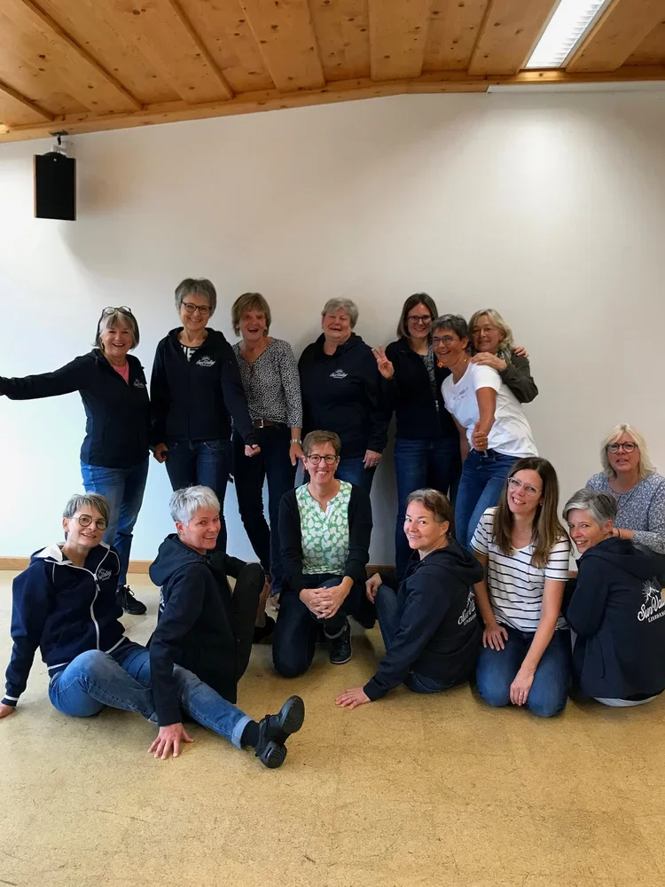 Gruppe von zwölf Frauen, die gemeinsam vor einer weißen Wand und in einem Raum mit Holzdecke stehen und sitzen, einige lächeln und posieren für das Foto.