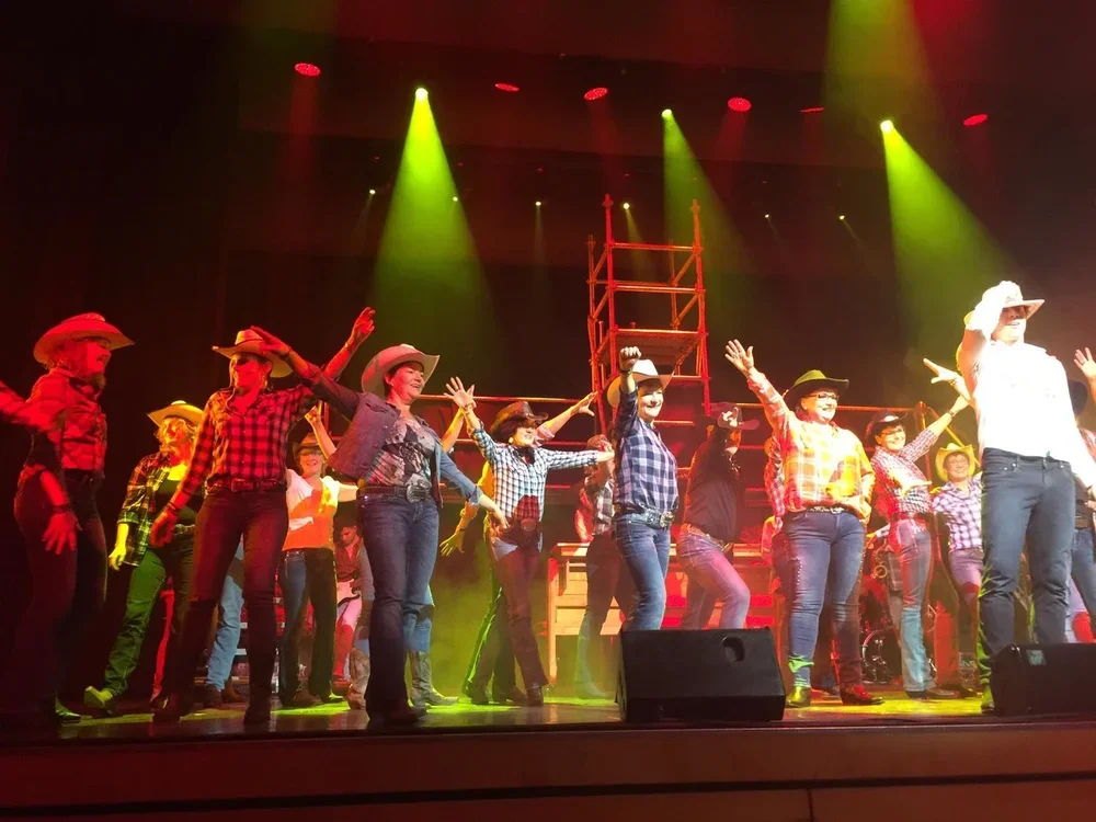 Bühne mit Darstellern in Cowboystimmung, viele Frauen in karierten Hemden und Cowboyhüten, rote und gelbe Bühnenlichter, Tanzaufführung im Theater.